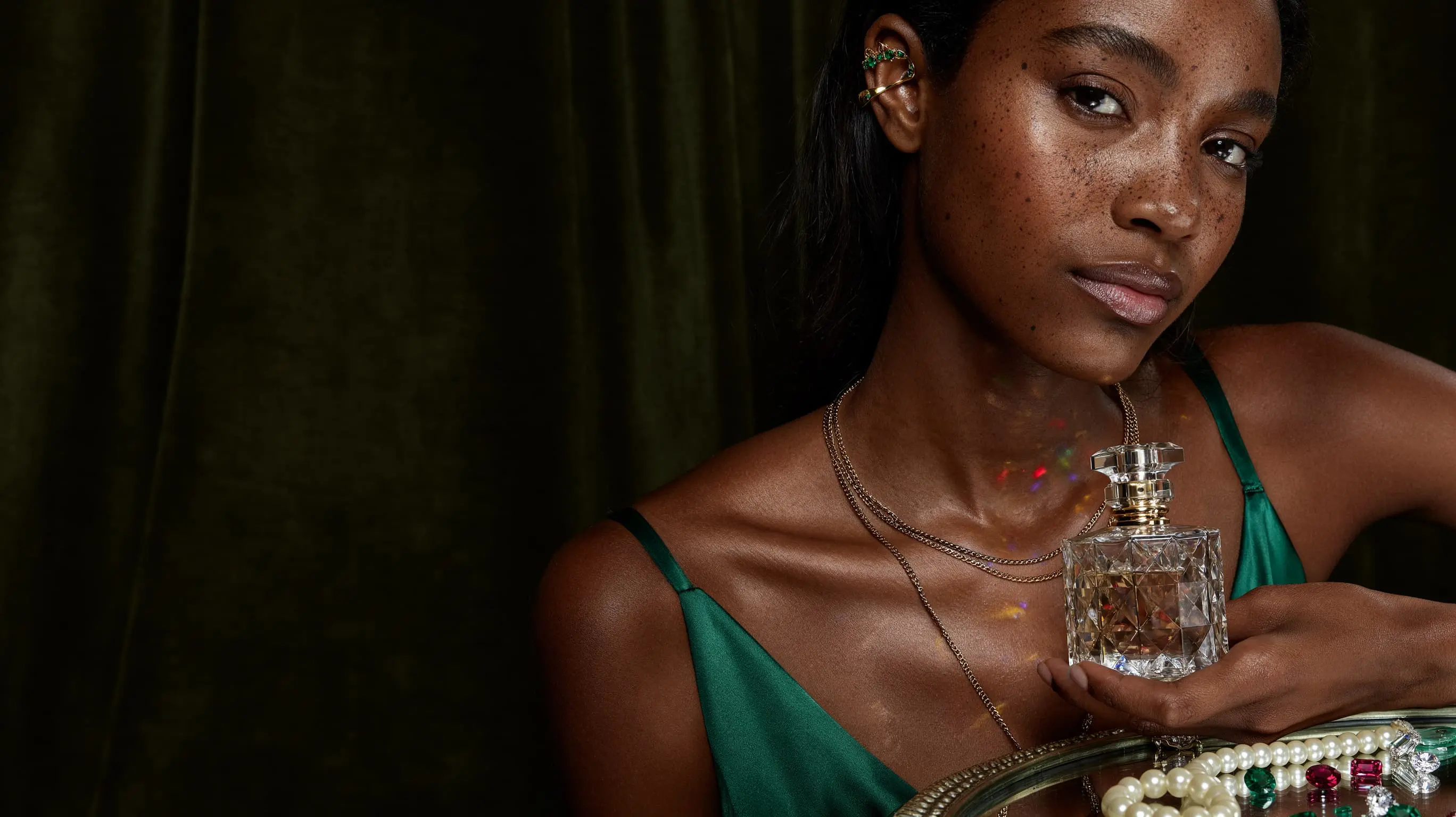 A beauty photograph of a woman in a green dress holding a crystal perfume bottle, jewelry in the foreground