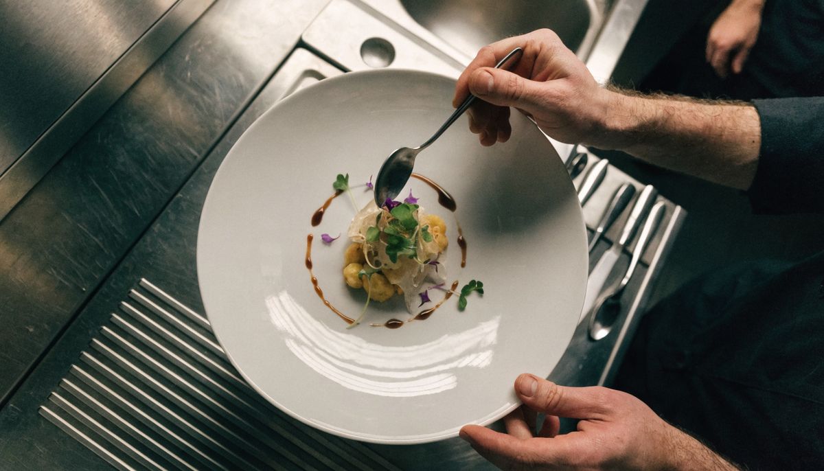 Top-down view of professional chef hands plating a fine dining dish on a white ceramic plate