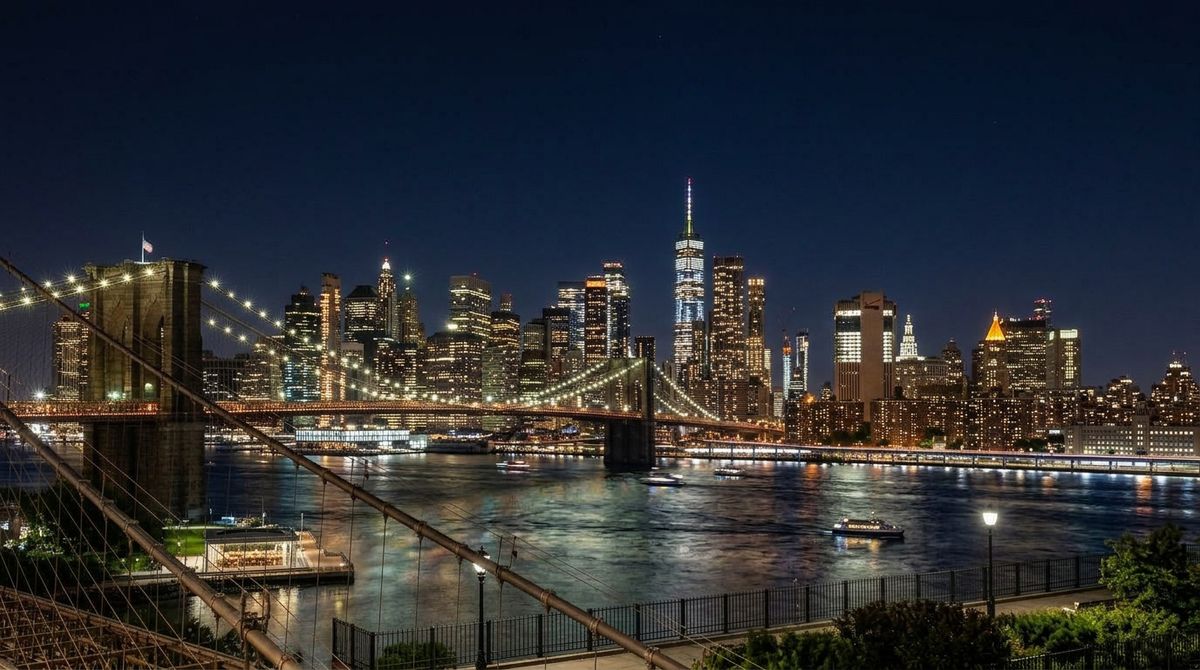 The same skyline view of Manhattan at night with thousands of office window lights glowing across the towers and the Brooklyn Bridge cables illuminated