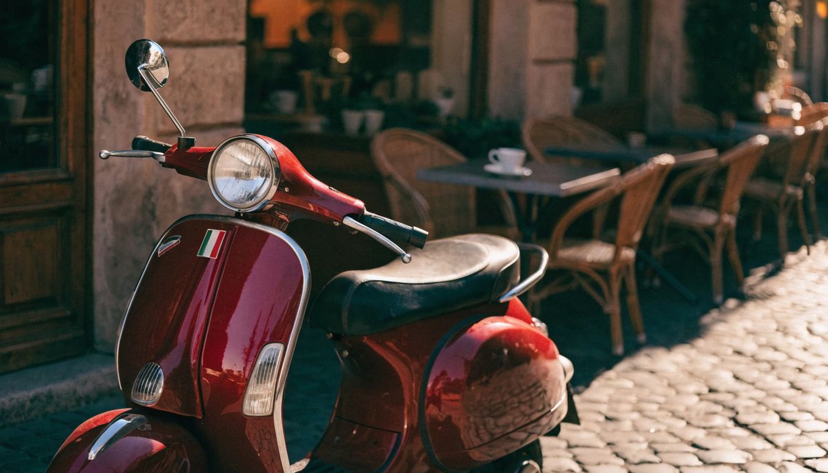 A vintage cherry red Vespa scooter parked on cobblestones outside a Roman cafe