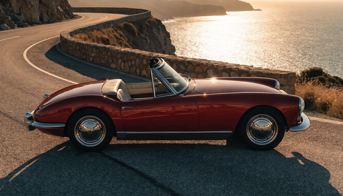 A vintage cherry red 1960s convertible parked side-on on a coastal cliff road at golden hour with the Mediterranean stretching below