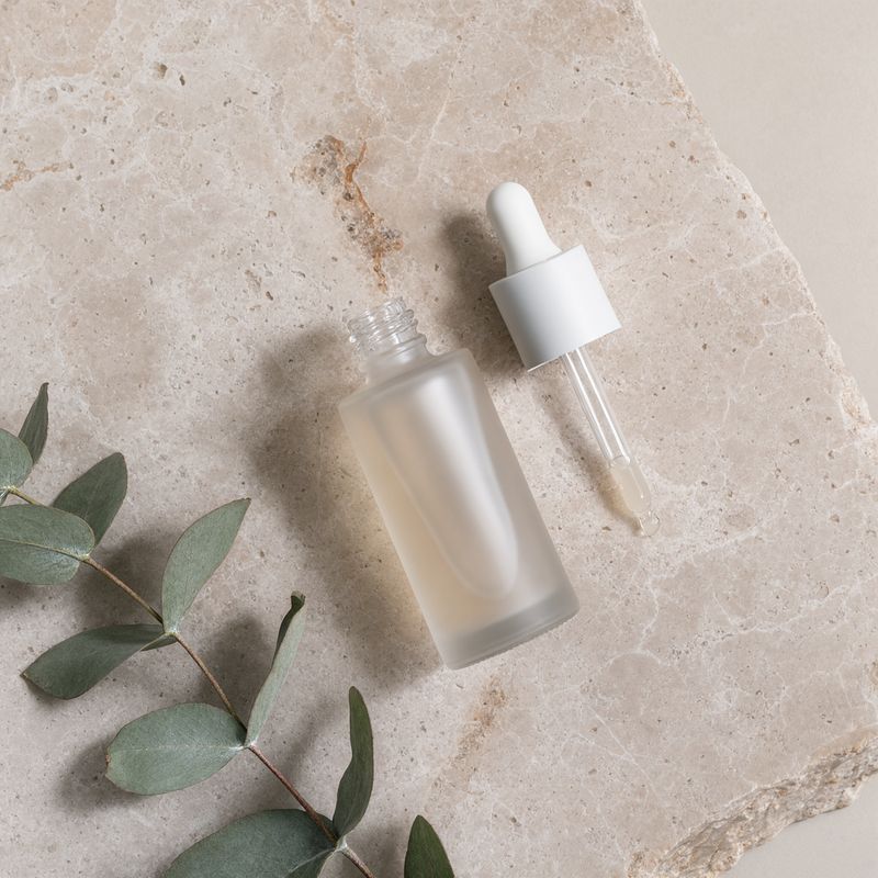 The same serum bottle lying on a travertine slab shot from directly above with a sprig of eucalyptus beside it