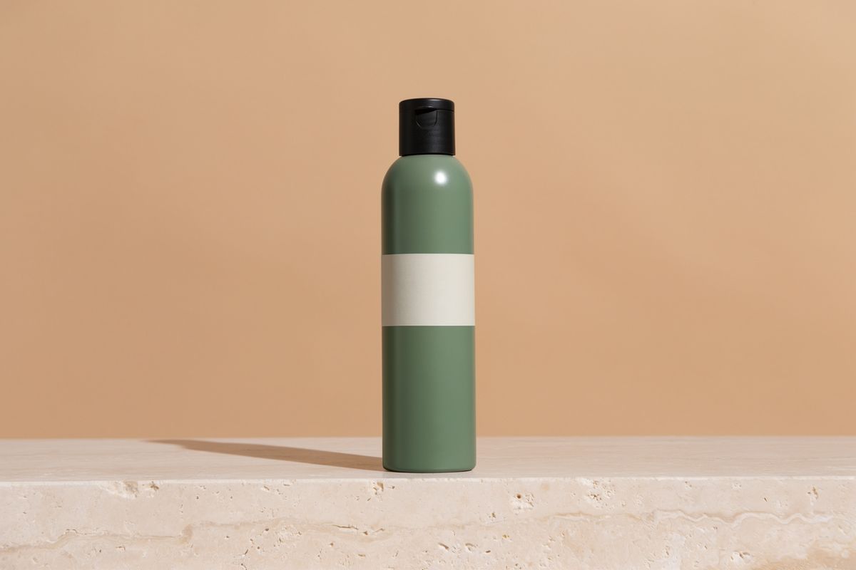 The sage green bottle on a travertine plinth against a warm sand backdrop with a hard shadow