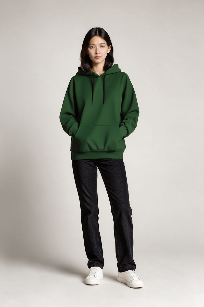 The same woman in the same pose wearing the same hoodie in deep forest green, with the jeans, sneakers and backdrop unchanged