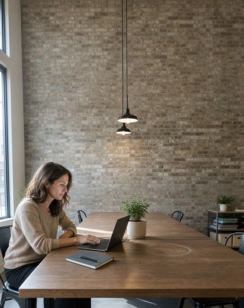 The same woman and workspace recomposed as a vertical frame with the brick wall and pendant lamps extending above her