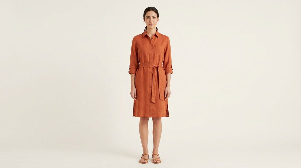 A full-length studio shot of a model wearing the same rust-orange linen shirt dress with the belt tied, standing against a bone-white backdrop
