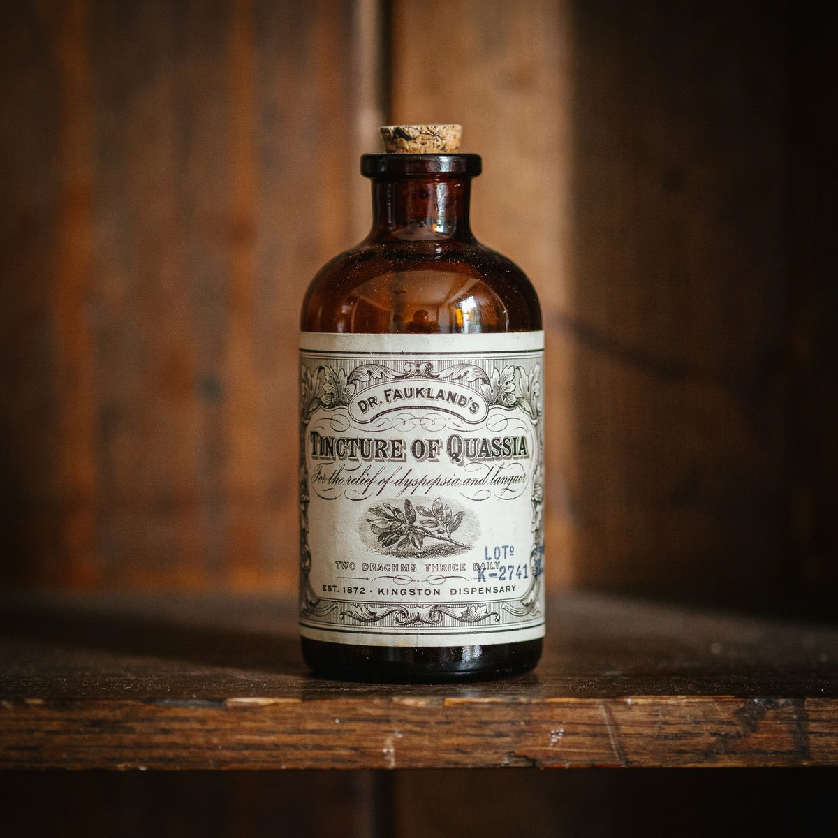 An ornate vintage apothecary label wrapped around a tall amber glass medicine bottle on a dark walnut shelf, reading 'Dr. Faukland's Tincture of Quassia' with dosage instructions and a manufacturer mark