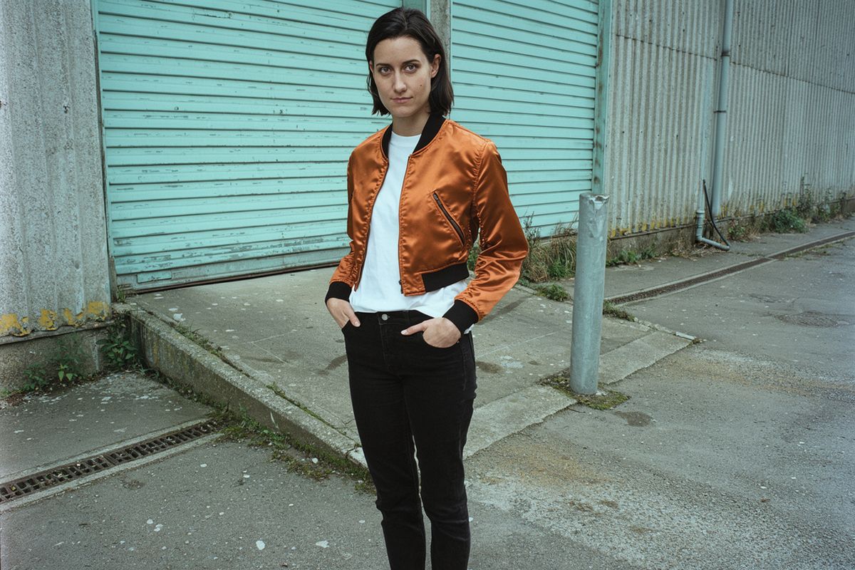 A woman in a burnt orange satin bomber jacket standing in front of pale teal warehouse shutters under overcast light
