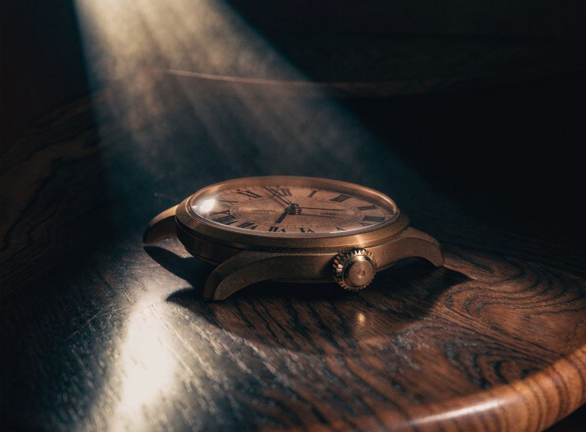 Dramatic chiaroscuro product photograph of a vintage brass mechanical wristwatch lying face-up on a dark walnut surface, a single warm directional light from the upper left creating a bright specular highlight on the domed crystal and casting a deep shadow behind the watch, dark chocolate background falling to black at the edges