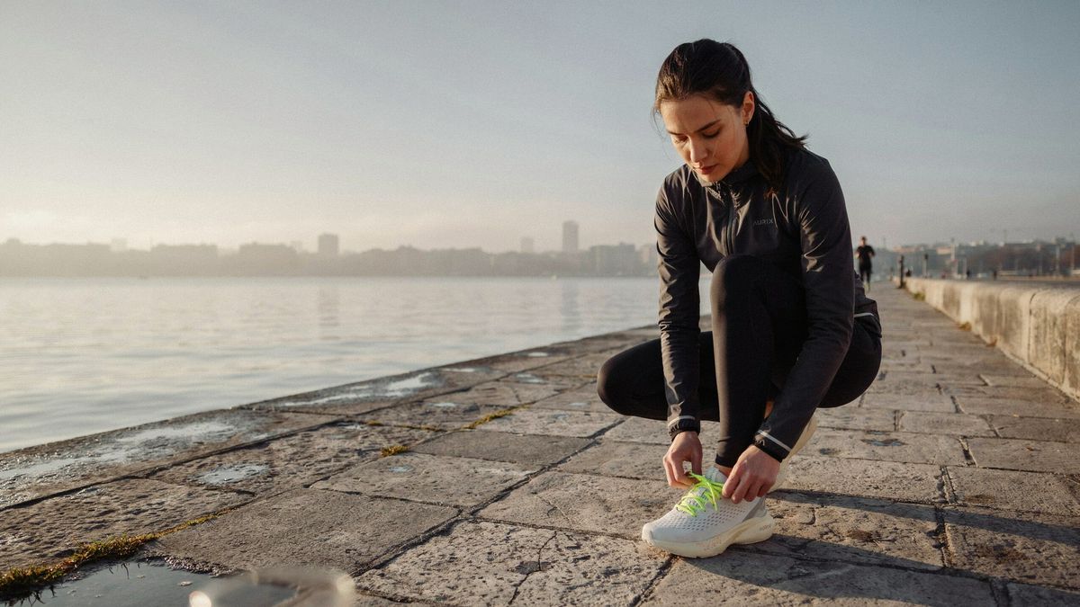 A woman in a charcoal running jacket crouching to tie a white running shoe on a stone waterfront at sunrise, the hazy city skyline across the water behind her