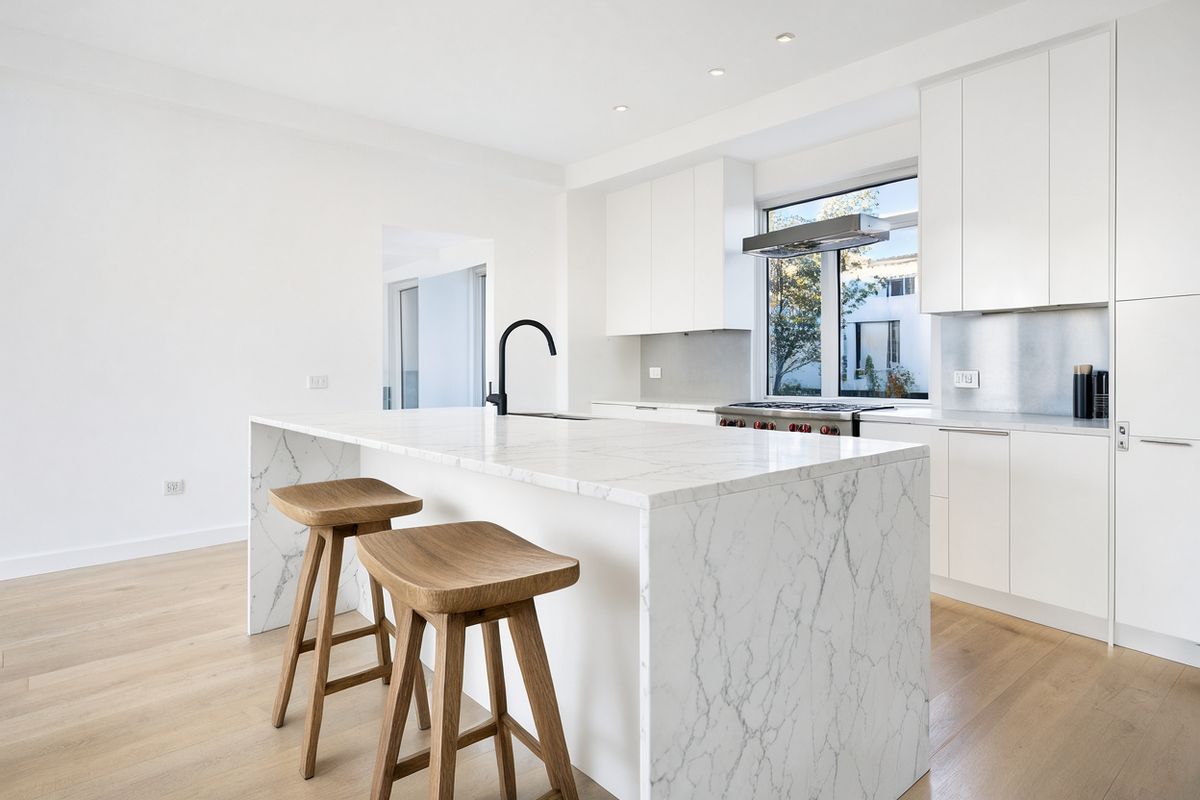 A white kitchen with a marble island, two oak stools and a window over the sink, the counters bare