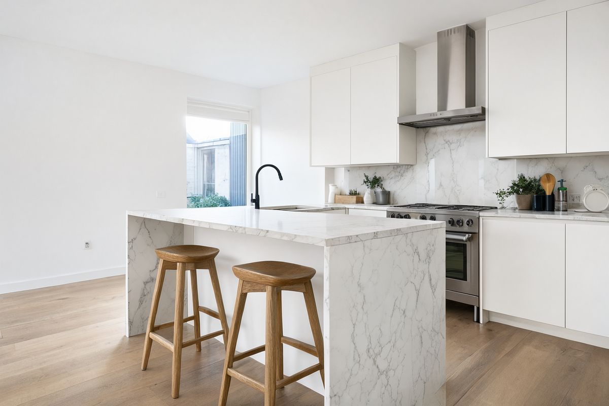 A white kitchen with a marble island and two oak stools, with potted herbs, a cutting board, canisters and bowls arranged along the back counter