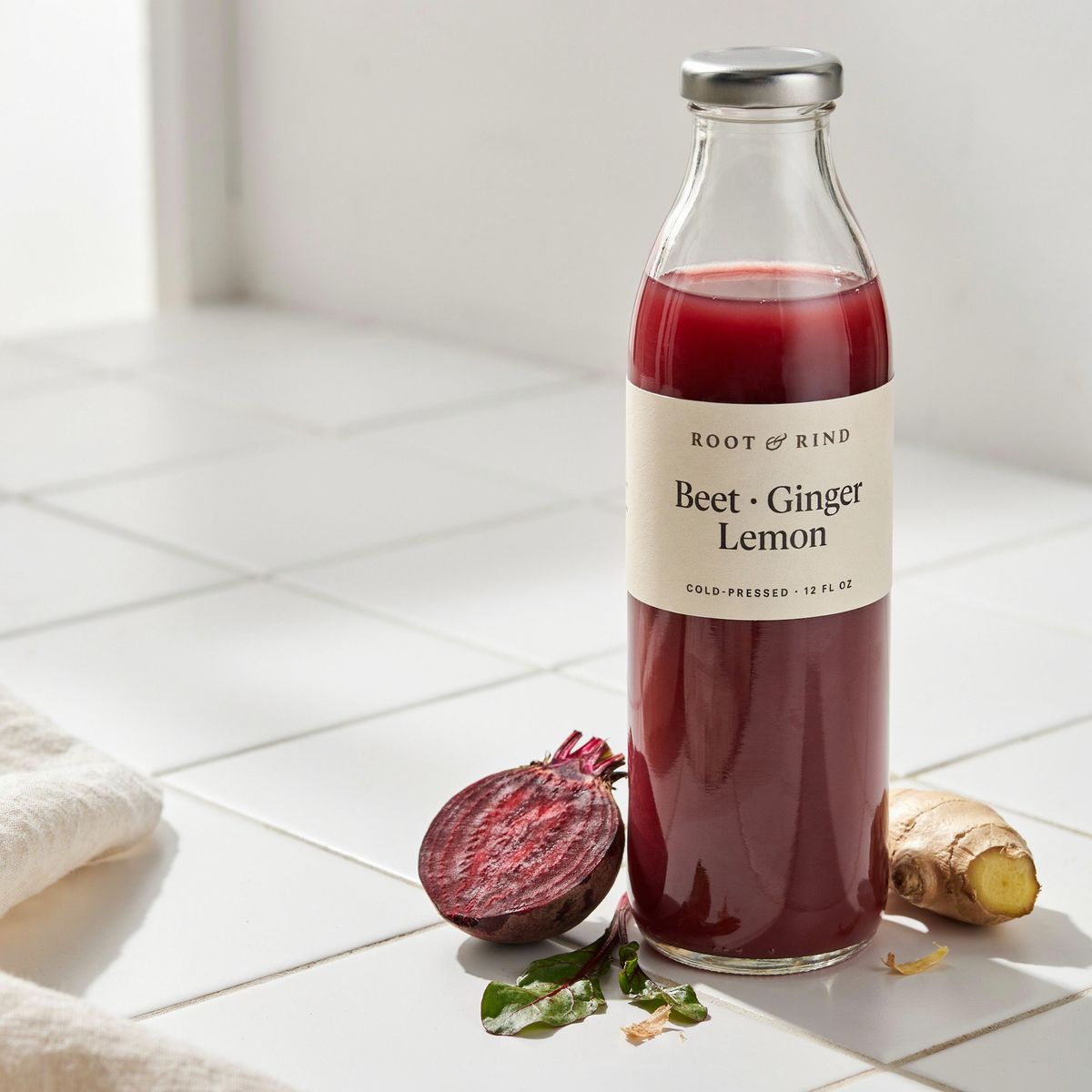 A glass bottle of magenta juice with a cream label reading ROOT & RIND at the top, Beet · Ginger and Lemon on two lines in a large serif, and COLD-PRESSED · 12 FL OZ in tiny capitals, beside a halved beet and ginger