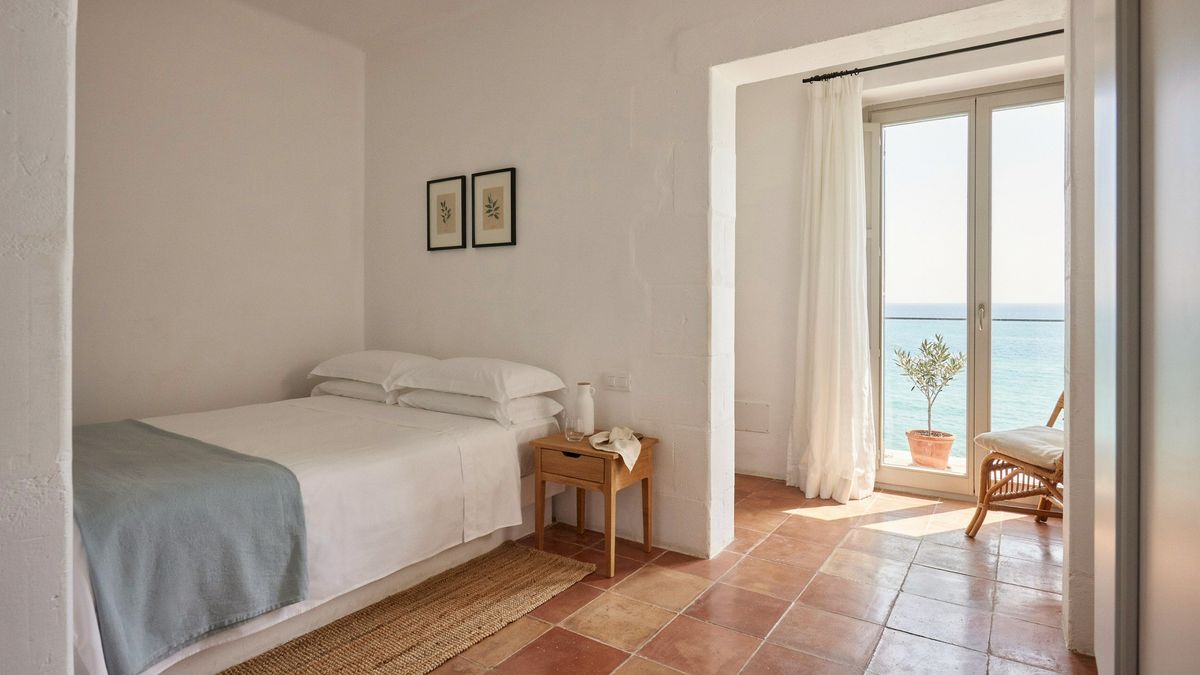 The detailed hotel room with the bed on the left under two prints, a rattan chair by the open doors, and an added olive tree on the balcony and jute rug by the bed