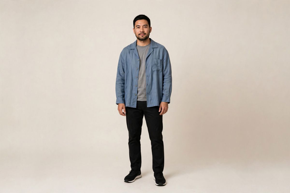 A full-length studio photo of a man in a dusty blue linen overshirt worn open over a grey t-shirt and black trousers against a warm-white backdrop