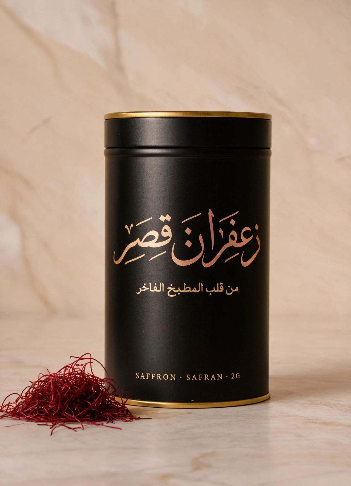 An Arabic luxury saffron packaging shot with a tall matte black saffron tin with a fine gold rim on a warm cream marble backdrop, the brand name زعفران قصر in warm rose gold thuluth calligraphy across the middle, a naskh tagline below, and a small SAFFRON · SAFRAN · 2G Latin label near the base