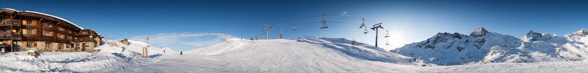 A panoramic strip of a ski resort: a snow-roofed timber lodge on the far left, a groomed piste with distant skiers in the center, a chairlift climbing toward a ridge, and snowy peaks under a low sun on the far right