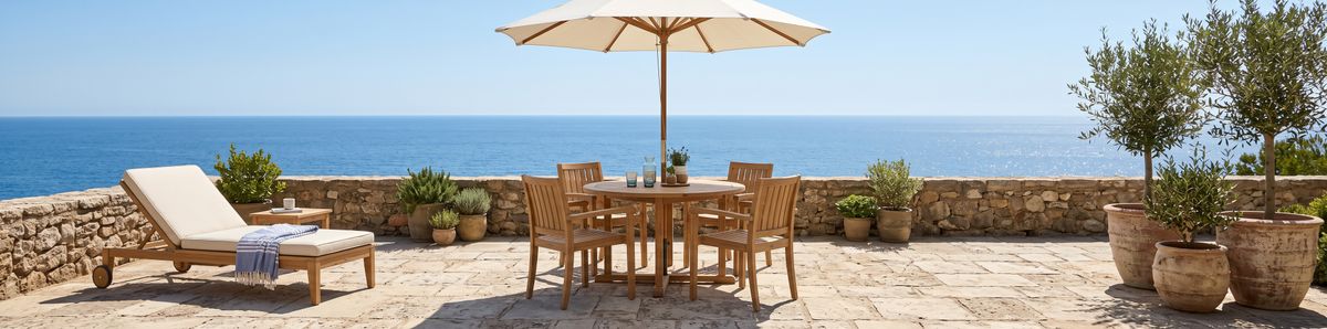 A wide banner of a sunlit stone terrace by the sea with a round dining table with four chairs under a parasol in the middle, a teak lounge chair with a striped throw on the left, and olive trees in terracotta planters on the right, in front of a low stone wall that runs the whole width