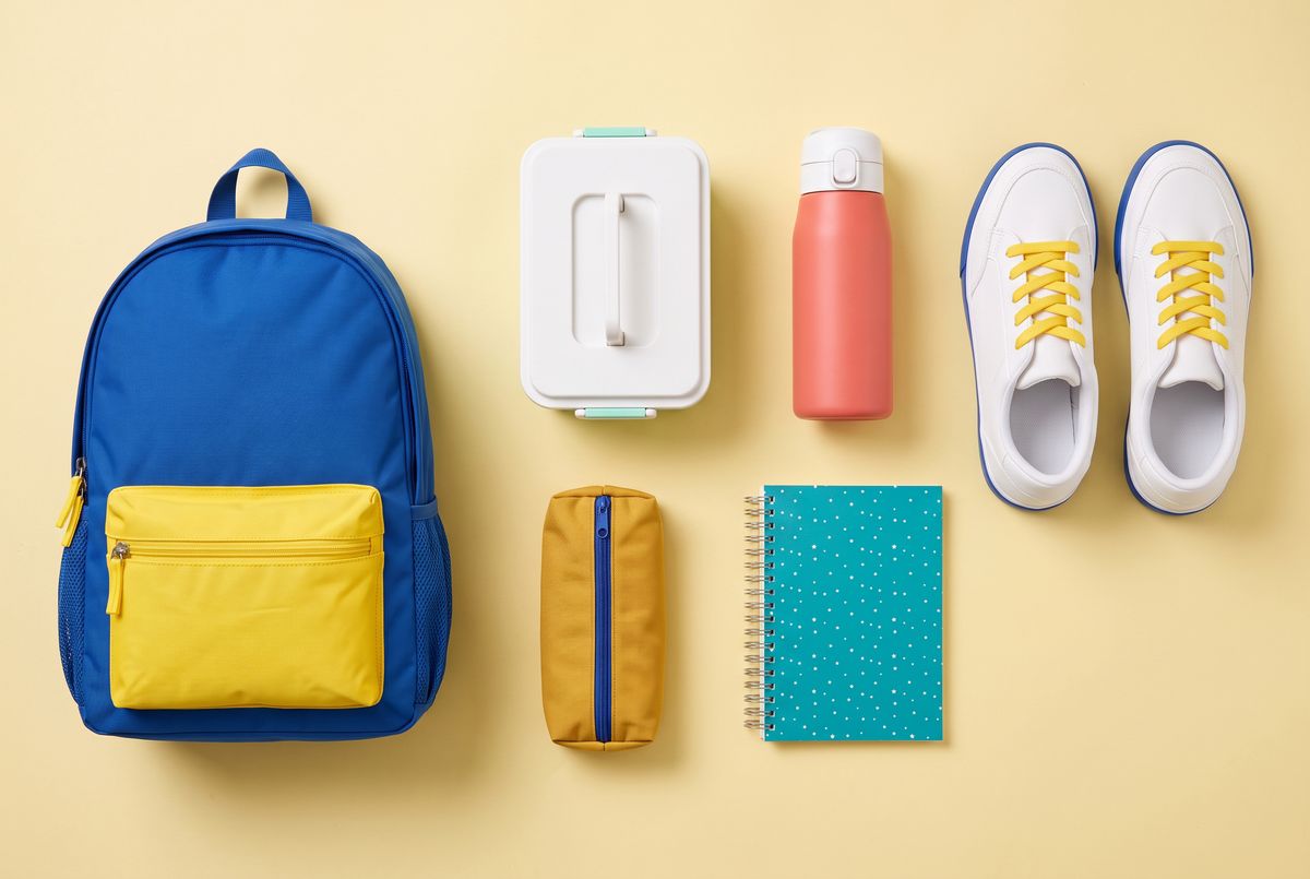 A flat-lay on pale yellow paper with a cobalt blue backpack on the left, a white-lidded lunch box with mint clips and a coral water bottle at upper center, a mustard pencil case and a teal star notebook at lower center, and white sneakers with blue soles on the right