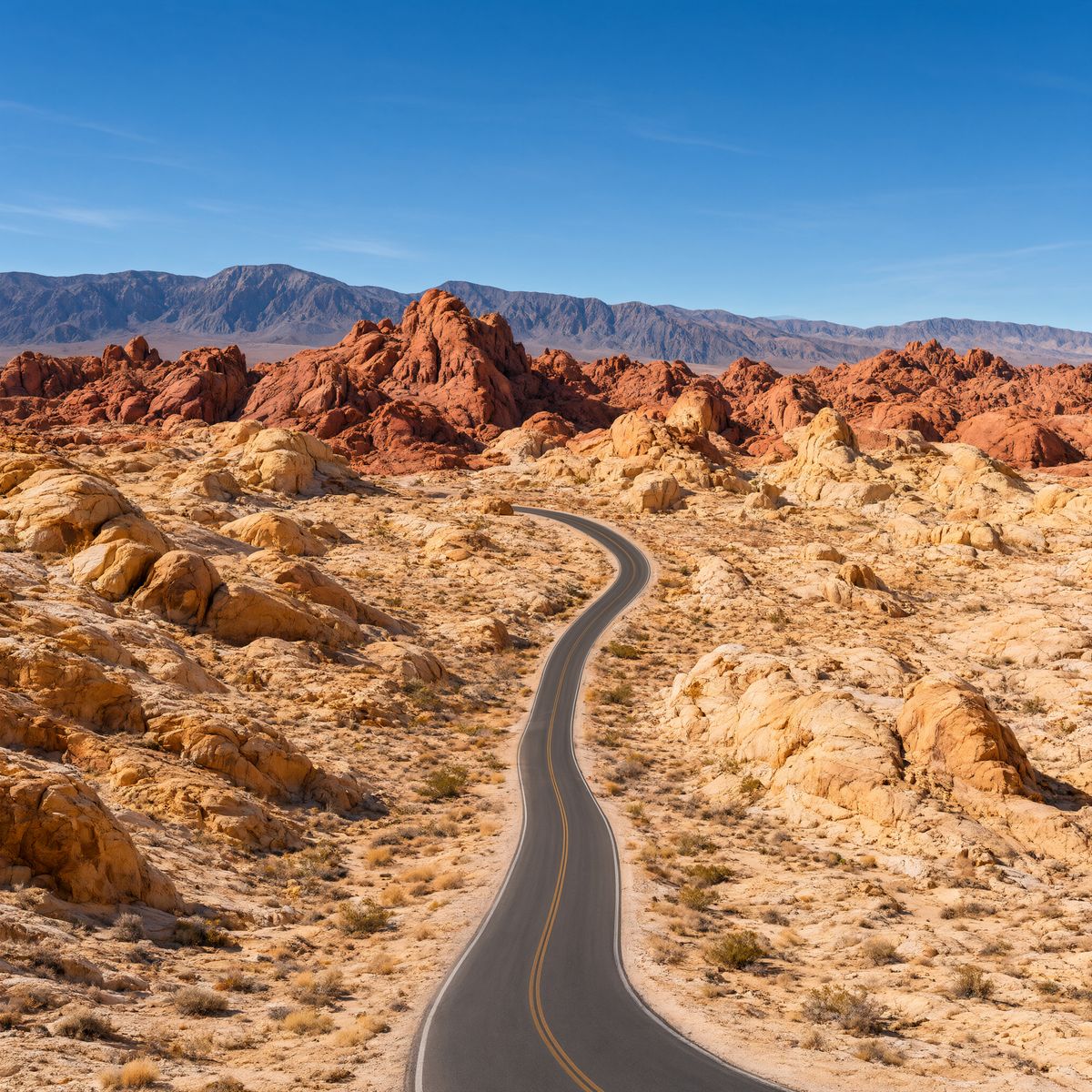 An elevated view of an empty two-lane road curving through red rock desert under a clear blue sky