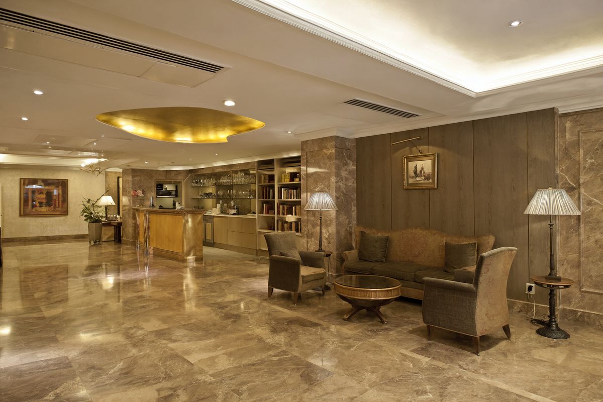 A hotel lobby with beige marble floors and walls, a wooden reception desk, a sofa with two armchairs, and a gold ceiling fixture