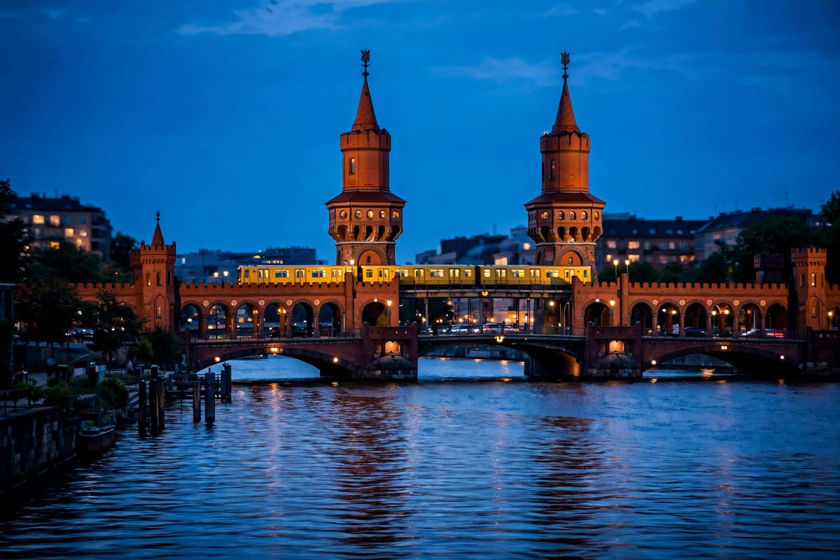 A lit brick river bridge with arches and towers photographed at dusk from a riverbank, a train crossing its upper level, a deep blue sky behind