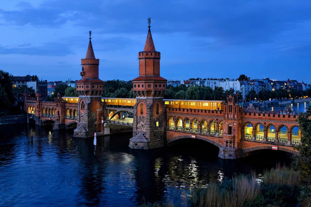 Berlin's Oberbaum Bridge at dusk with its twin stepped-gable brick towers, the arcaded lower road deck and the upper viaduct carrying a yellow U-Bahn train, reflected in the Spree