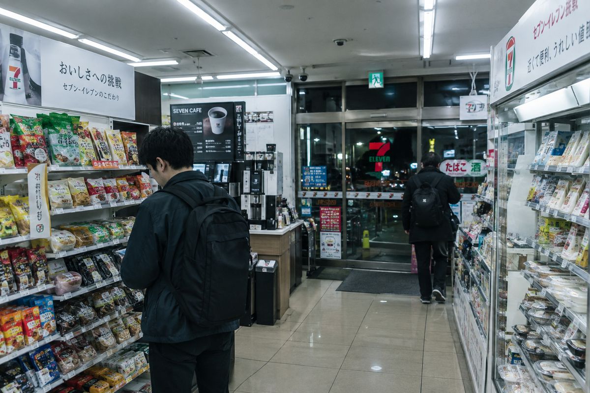 The interior of a Tokyo convenience store at night under flat fluorescent light, with packed shelves, a chilled drinks cabinet along the back wall and a hot food counter by the till