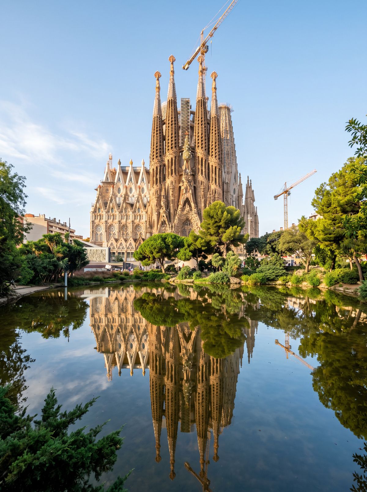 The Sagrada Família in Barcelona seen across a pond on a clear morning, with two construction cranes over an unfinished center, its spires reflected in the water
