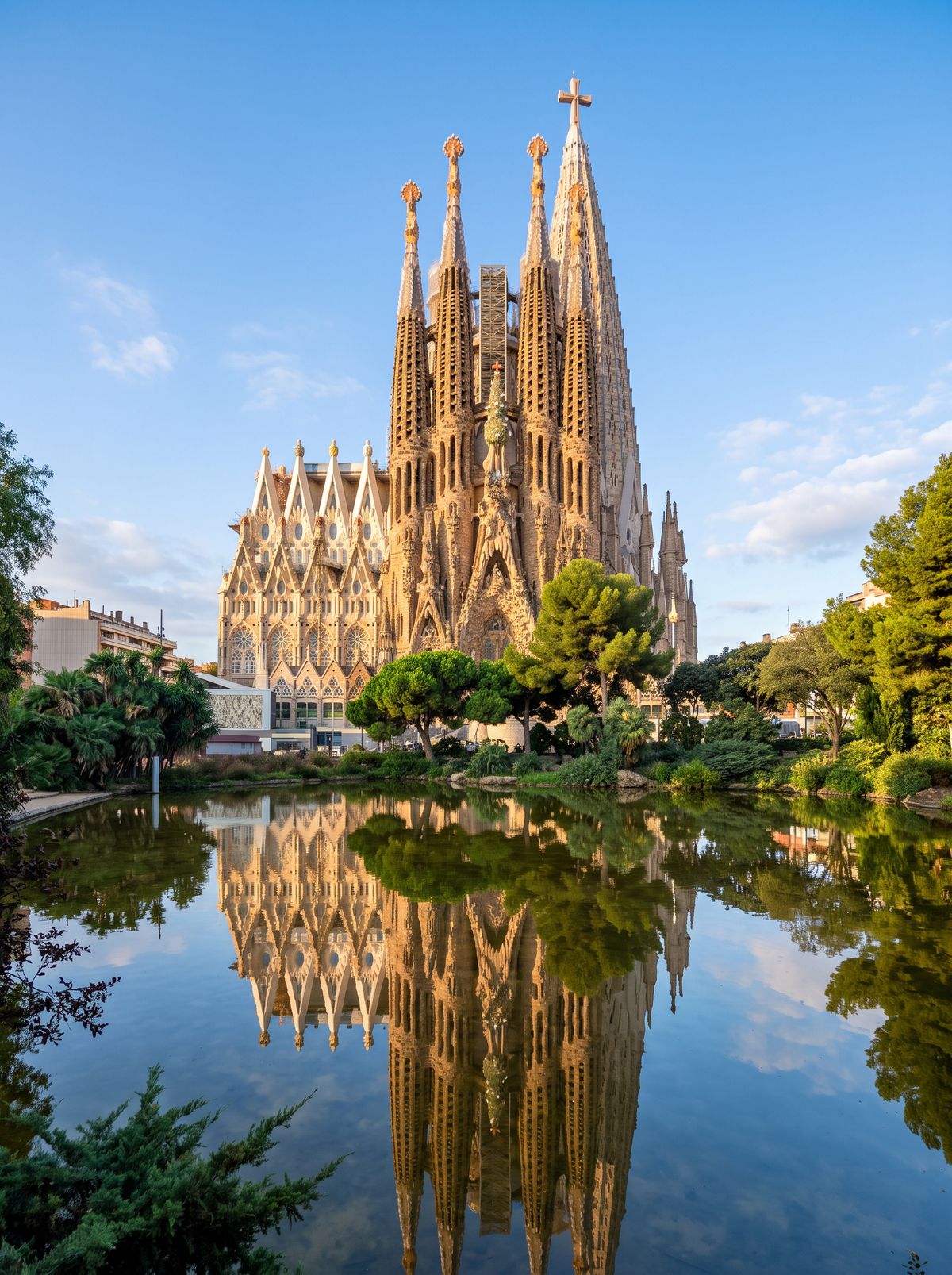 The Sagrada Família in Barcelona seen across a pond on a clear morning, with no cranes and the completed central tower and its cross rising above the other spires, reflected in the water
