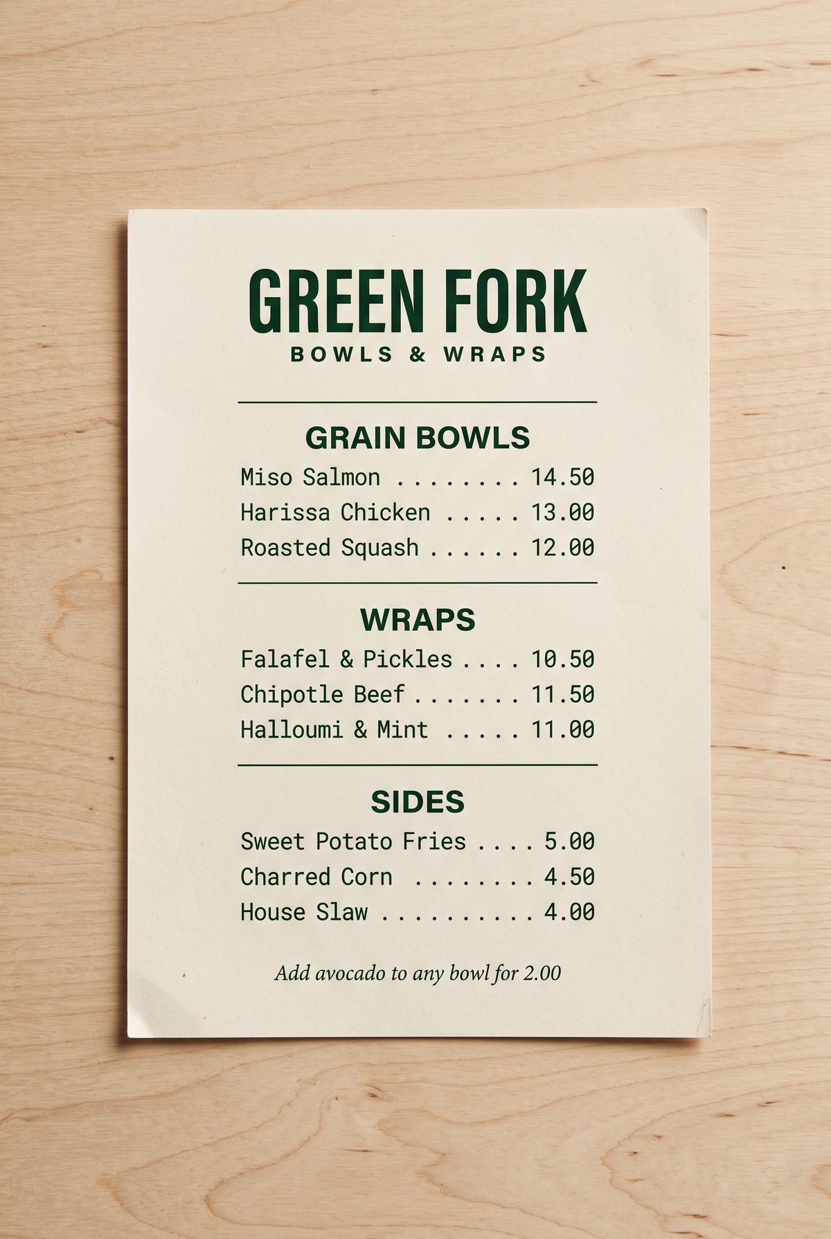 A cream takeaway menu with dark green type on a wood counter, headed GREEN FORK, BOWLS & WRAPS, with three ruled sections, GRAIN BOWLS, WRAPS, and SIDES, each listing three dishes with prices aligned on the right, and a line about adding avocado at the bottom