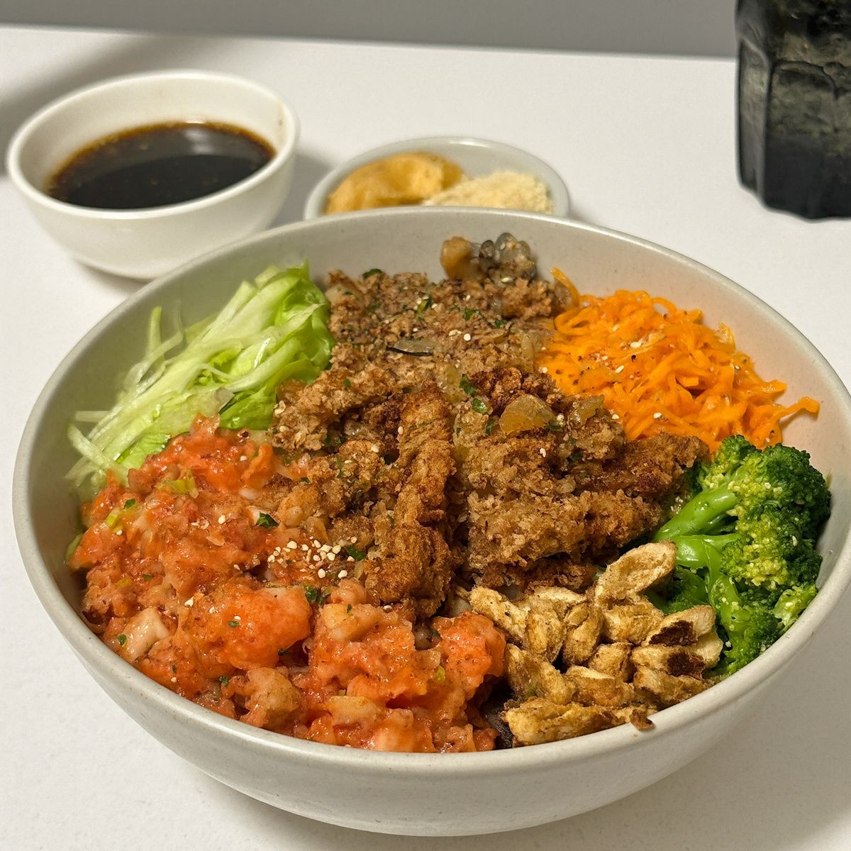 A ceramic bowl of shredded lettuce, carrot, broccoli, chopped tomato, and fried toppings seen from a three-quarter angle, with small dishes of soy sauce and ginger behind it