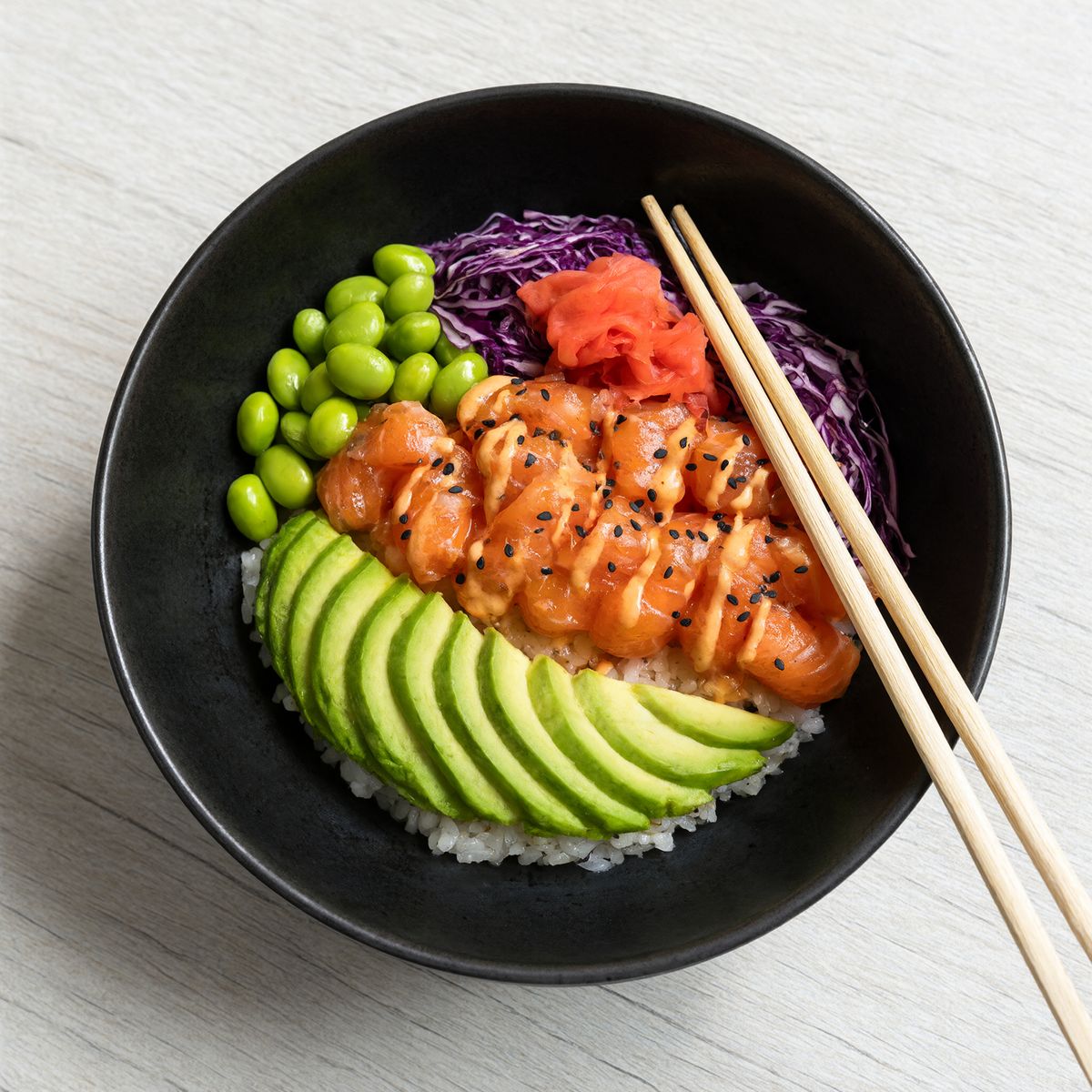An overhead shot of a black ceramic poke bowl on pale wood with salmon cubes under sesame seeds and a mayo drizzle, fanned avocado, edamame, purple cabbage, and pickled ginger, with chopsticks across the right edge
