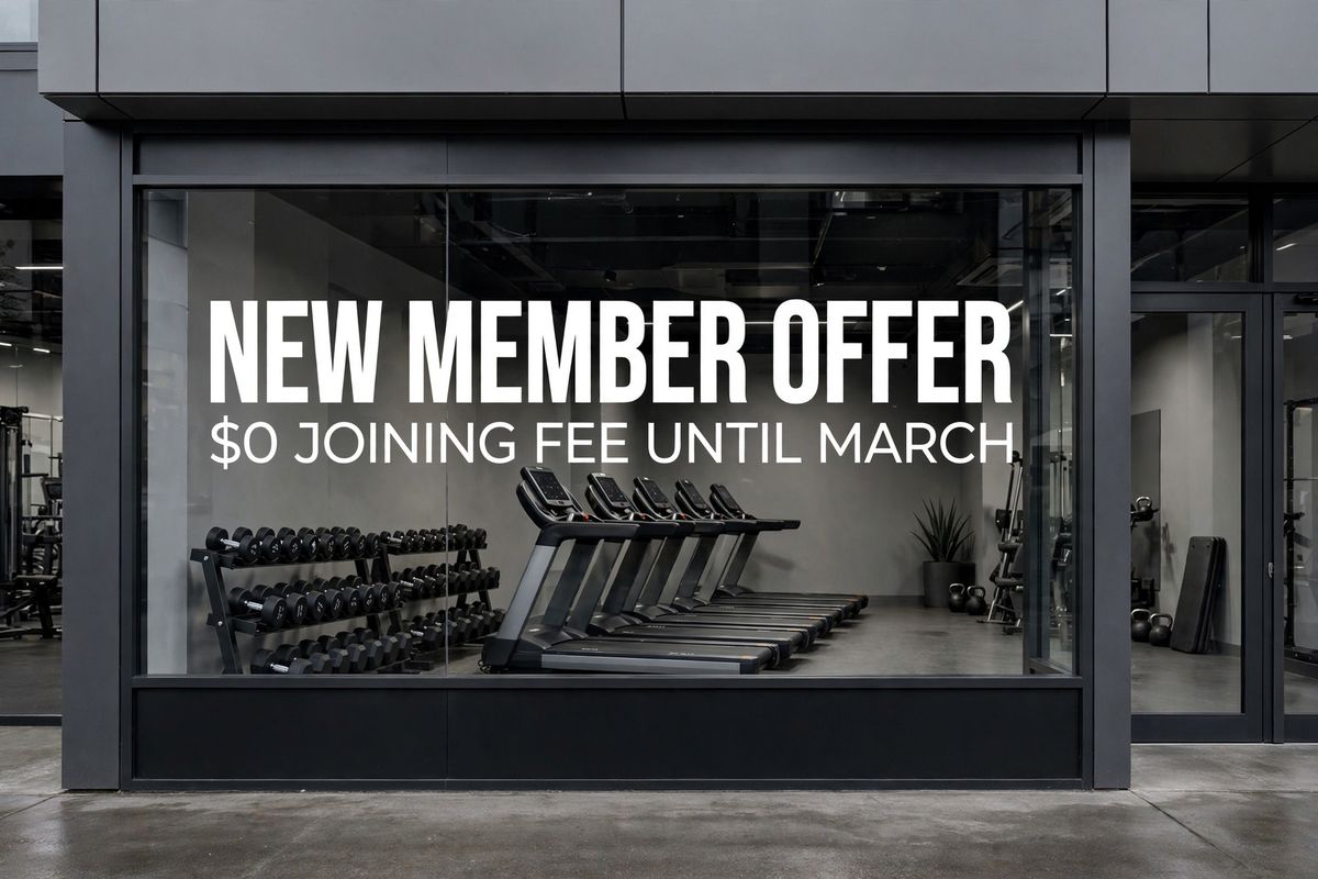 A gym storefront window carrying a white vinyl decal that reads NEW MEMBER OFFER in heavy condensed capitals above $0 JOINING FEE UNTIL MARCH in a lighter weight, with dumbbell racks and treadmills visible through the glass