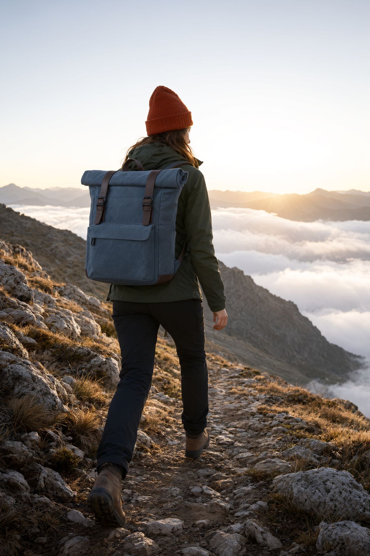 A hiker in a rust beanie and green jacket climbing a ridge above a sea of cloud at sunrise, wearing the slate blue backpack