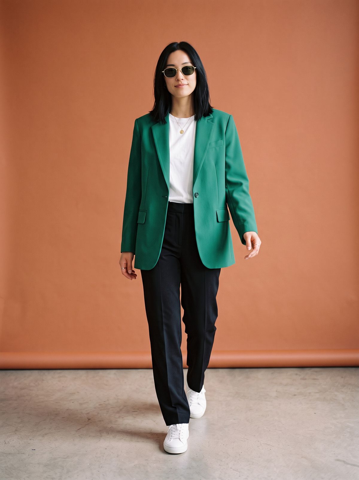 The same woman in the same stride wearing an emerald green blazer, black tailored trousers, and gold-rimmed sunglasses against a terracotta backdrop, with the white T-shirt and sneakers unchanged
