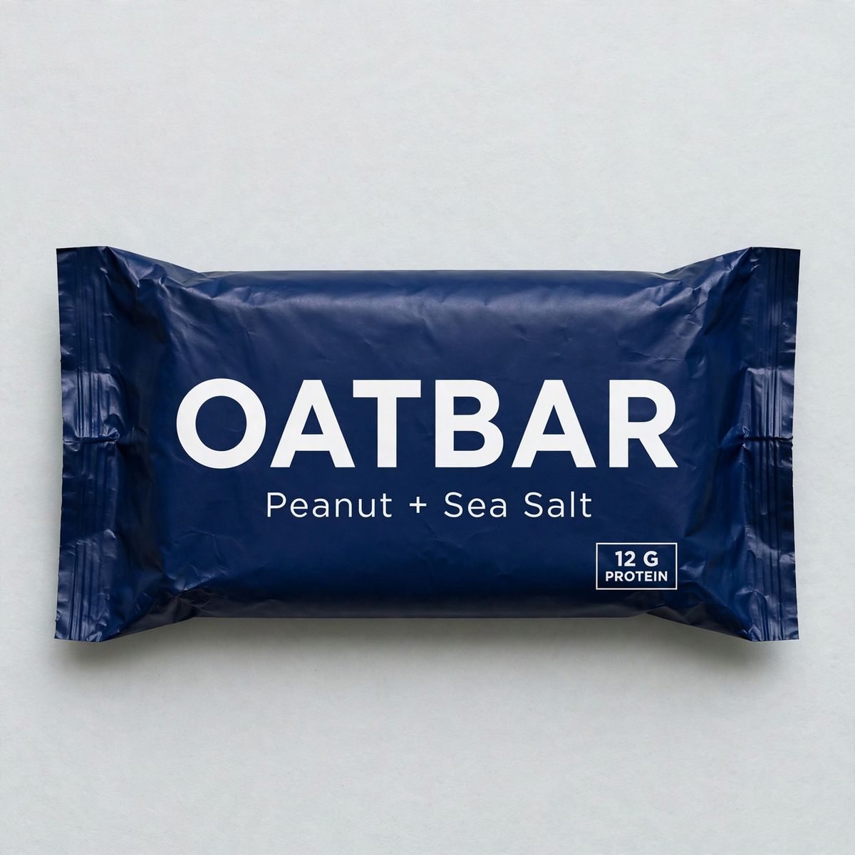 A flat lay of a deep navy protein bar wrapper printed with OATBAR in large white capitals, Peanut + Sea Salt beneath it, and 12 G PROTEIN in a small white box in the lower right corner