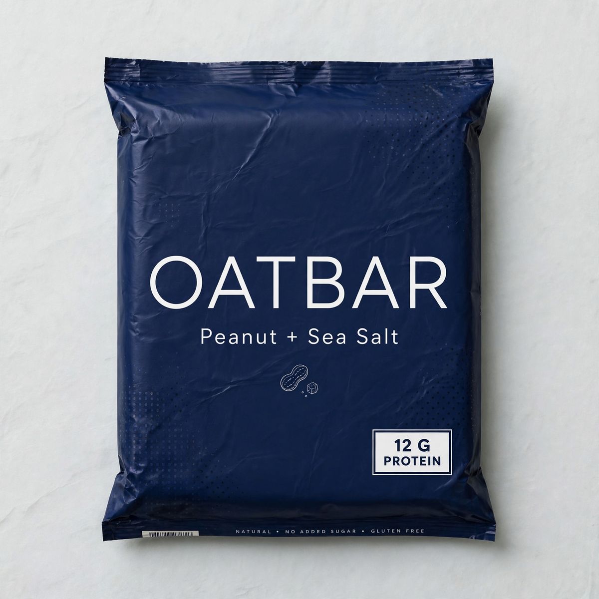 A flat lay of a deep navy protein bar wrapper printed with OATBAR in large white capitals, Peanut + Sea Salt beneath it, and 12 G PROTEIN in a small white box in the lower right corner