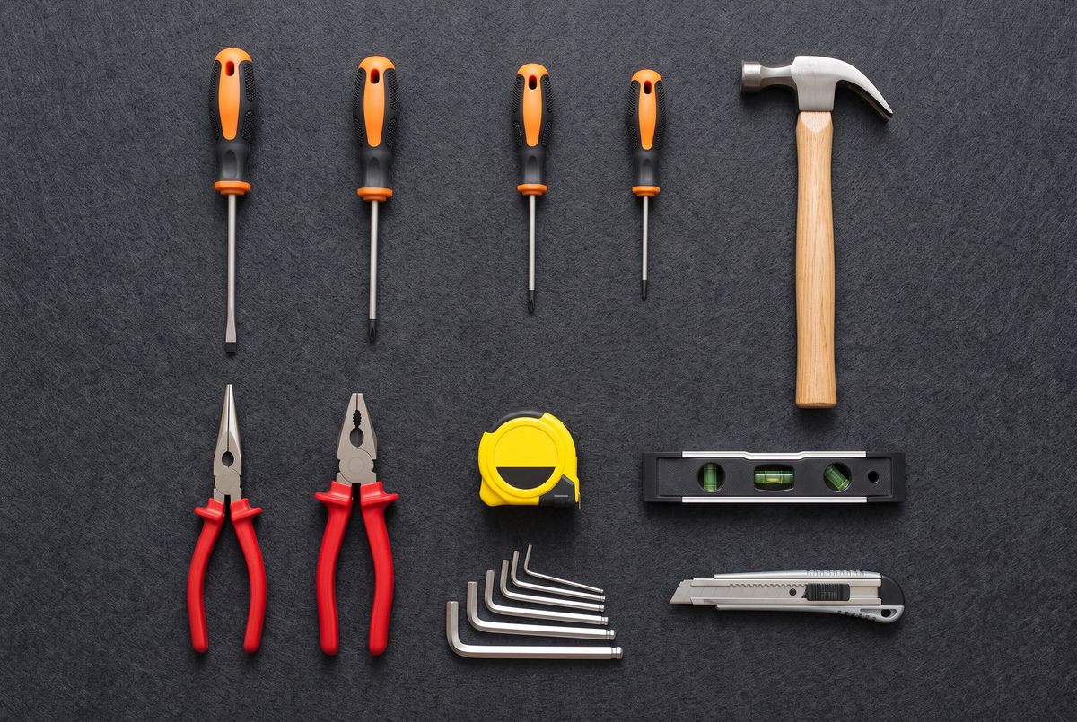 An overhead flat-lay of a home tool kit on dark gray felt in three rows: four upright screwdrivers with orange handles and a claw hammer, then two pliers with red grips, a yellow tape measure, and a torpedo level, then six nested hex keys and a utility knife