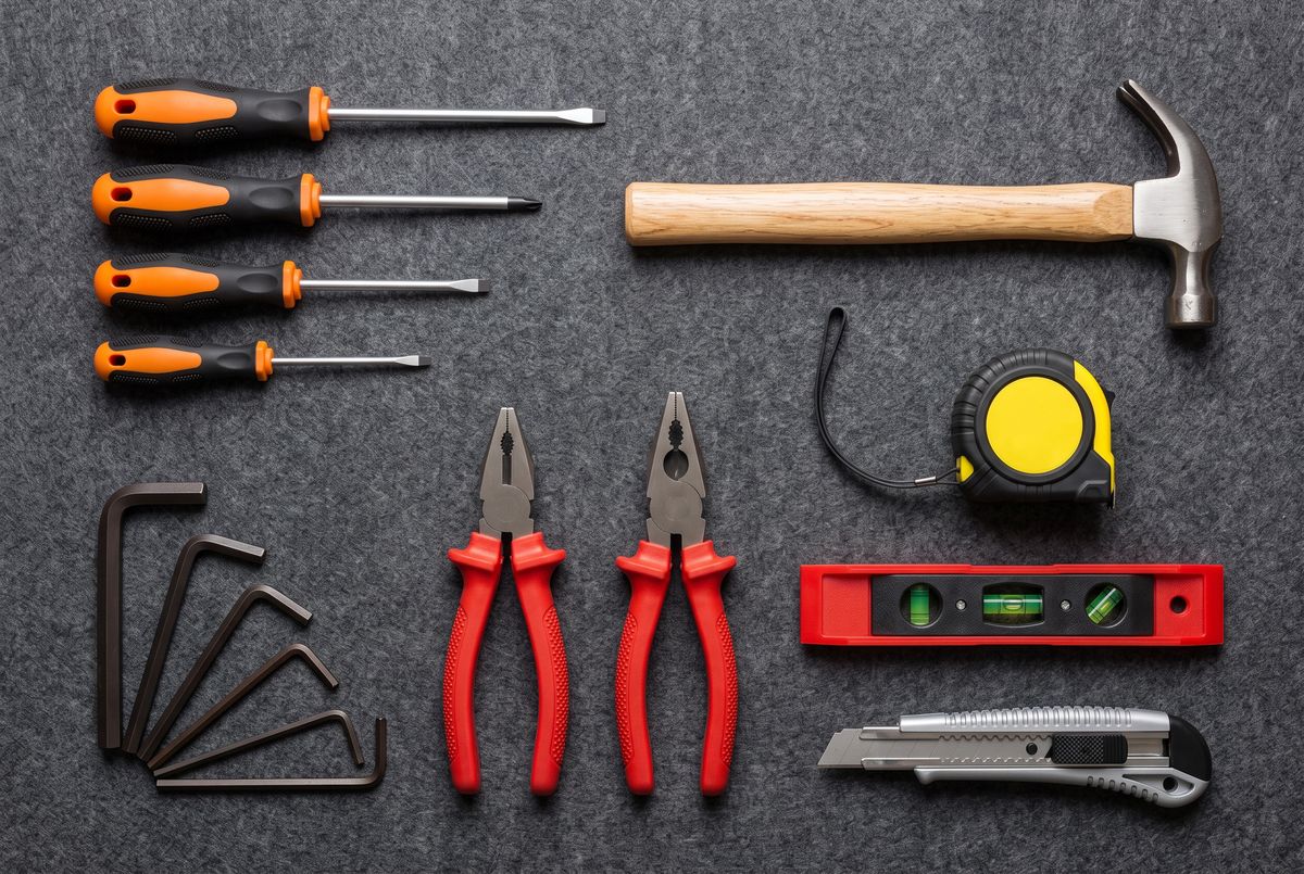 An overhead flat-lay of a home tool kit on dark gray felt: four screwdrivers with orange handles stacked at the top left, a claw hammer, two pliers with red grips, a yellow tape measure, a red torpedo level, a utility knife, and six dark hex keys fanned in a quarter circle at the bottom left