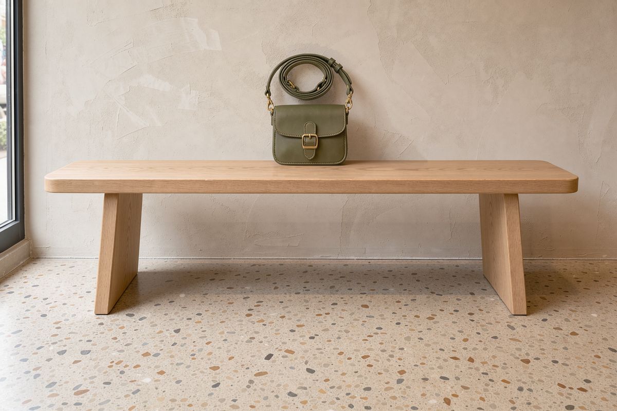 An olive green crossbody bag standing on a pale oak bench against a lime-washed wall