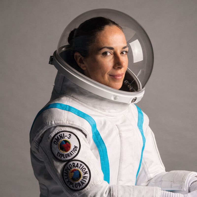 A female astronaut in a sleek white pressure suit with cyan accent stripes, a clear bubble helmet, and OMNI-3 EXPLORATION patches on her left arm