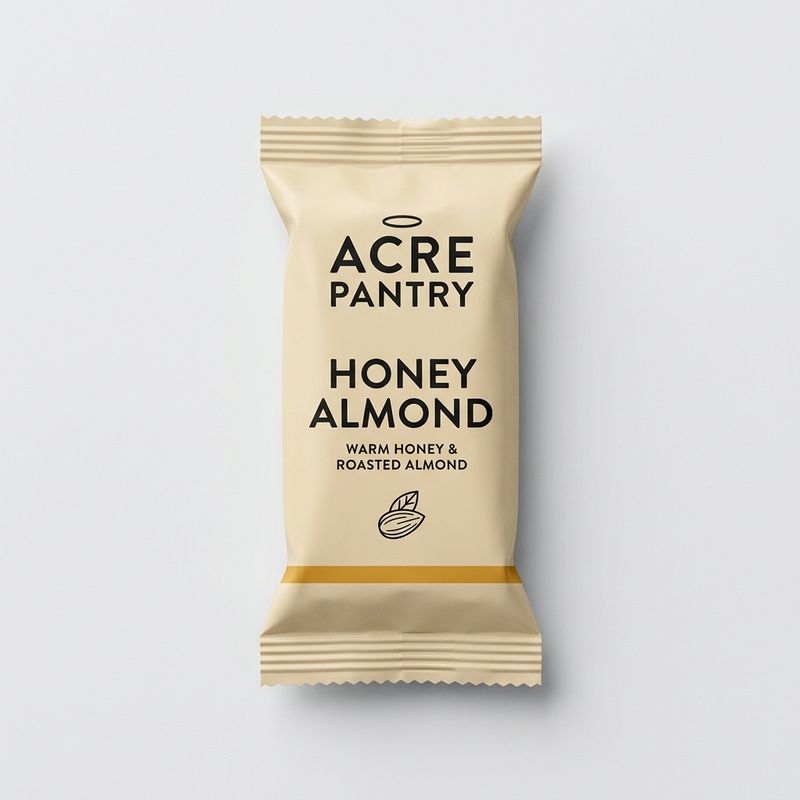 Flat-lay product photograph of the Acre Pantry Honey Almond granola bar in a flow-wrap pillow pouch, kraft cream paper with ACRE PANTRY wordmark, HONEY ALMOND flavor name, almond illustration, and amber gold accent stripe