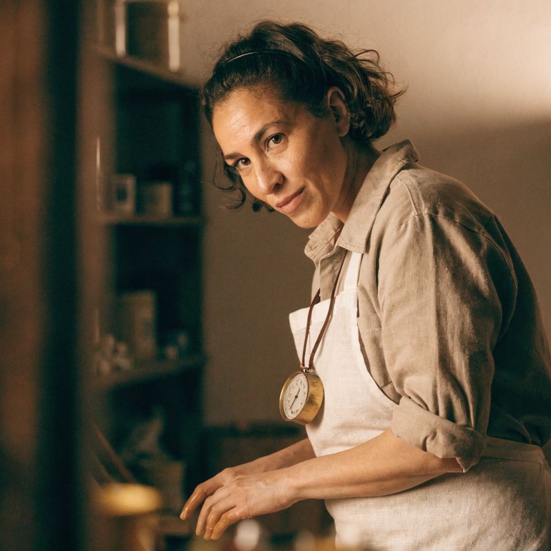 An artisan candle maker with short dark hair and a white linen apron, wearing a brass thermometer on a leather cord around her neck