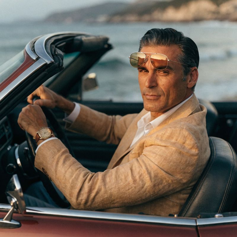 A male driver in his forties in a tan linen blazer with aviator sunglasses on his forehead and a vintage gold wristwatch