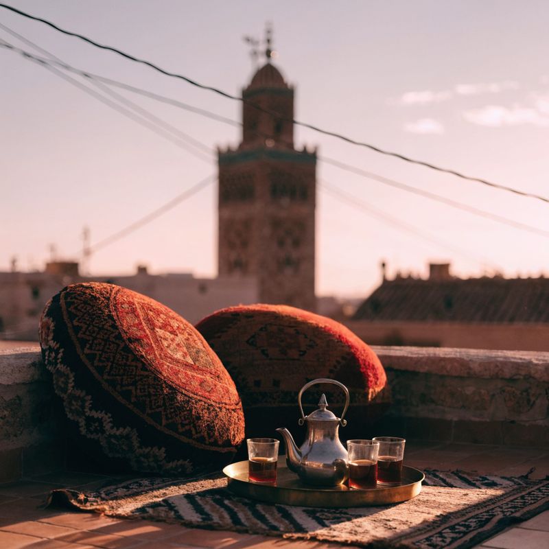 A Marrakech rooftop terrace at golden hour with a Berber rug, embroidered cushions, and the Koutoubia minaret silhouetted behind