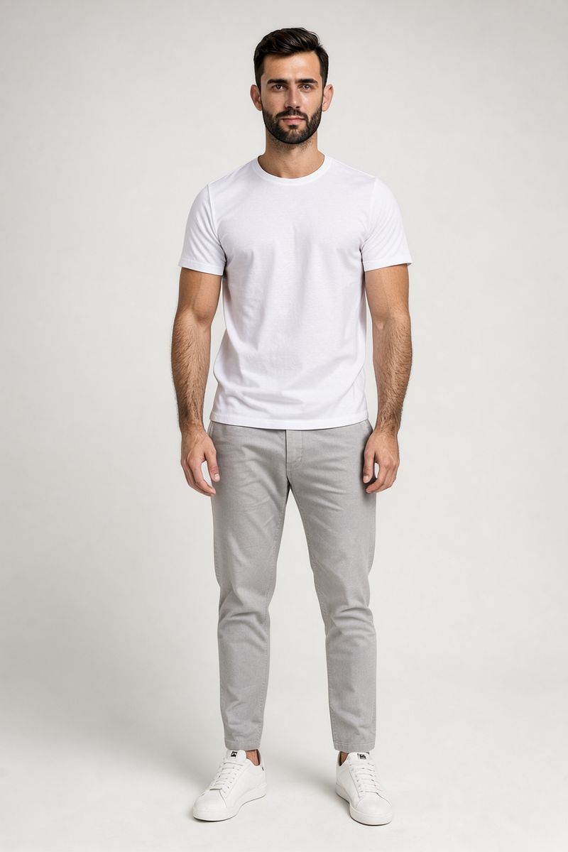 A full-length studio photo of a bearded man in a white t-shirt, light gray chinos and white sneakers on a gray backdrop