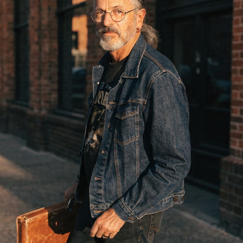 A music collector in his fifties with a salt-and-pepper ponytail, wire-frame glasses, and a denim jacket over a faded band tee, holding a leather record case