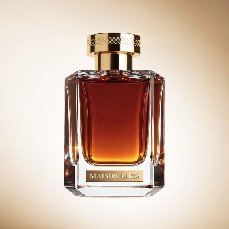 A luxury perfume bottle product shot: a tall faceted clear glass column filled with deep amber liquid, a polished gold cap, and an embossed MAISON LUNA label