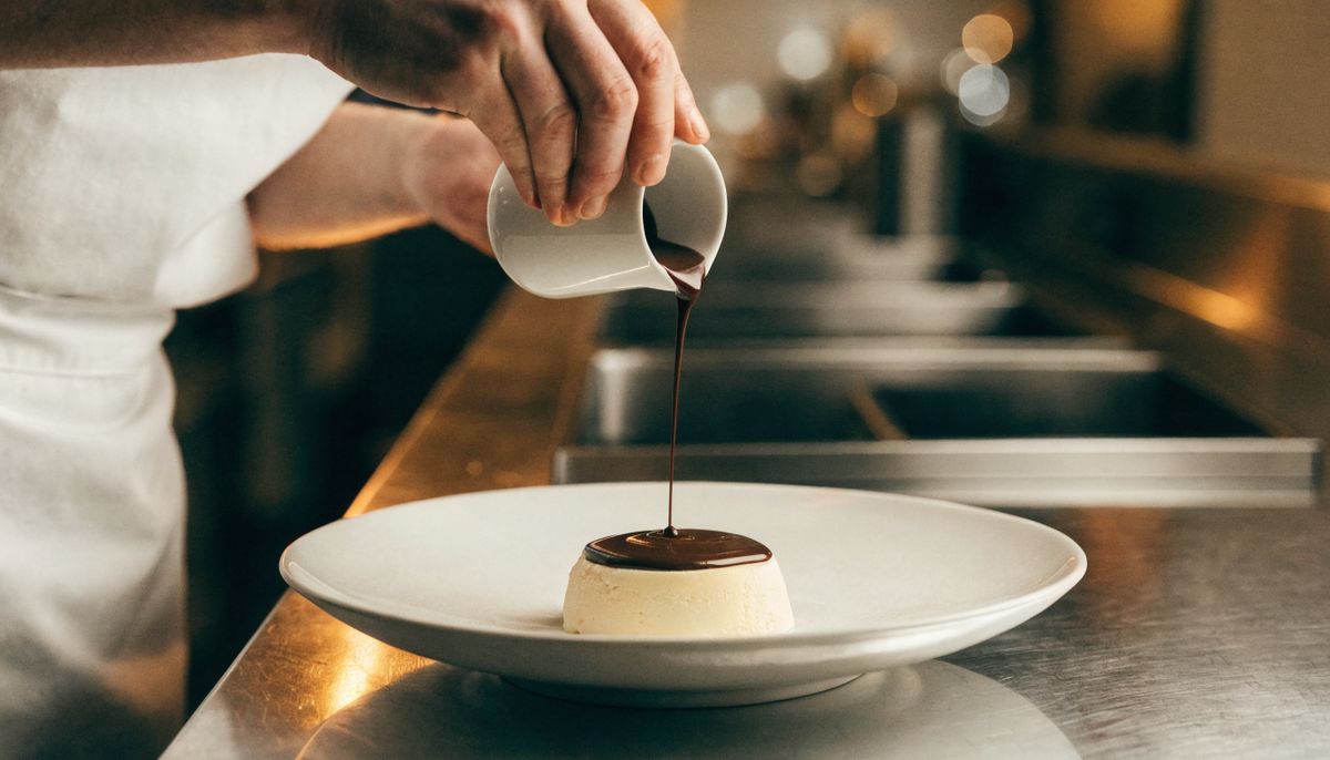A chef in a white apron pouring dark chocolate sauce from a ceramic pitcher over a vanilla panna cotta on a white plate